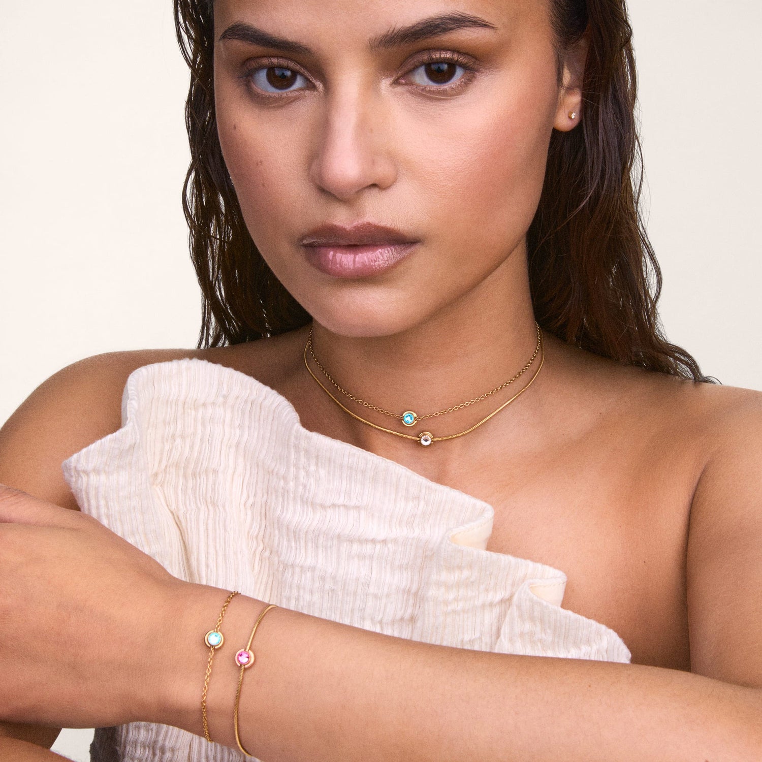 A woman wears the December Charm S925 Sterling Silver with a blue topaz-coloured crystal on a gold chain necklace and matching bracelet, styled with a cream textured top.