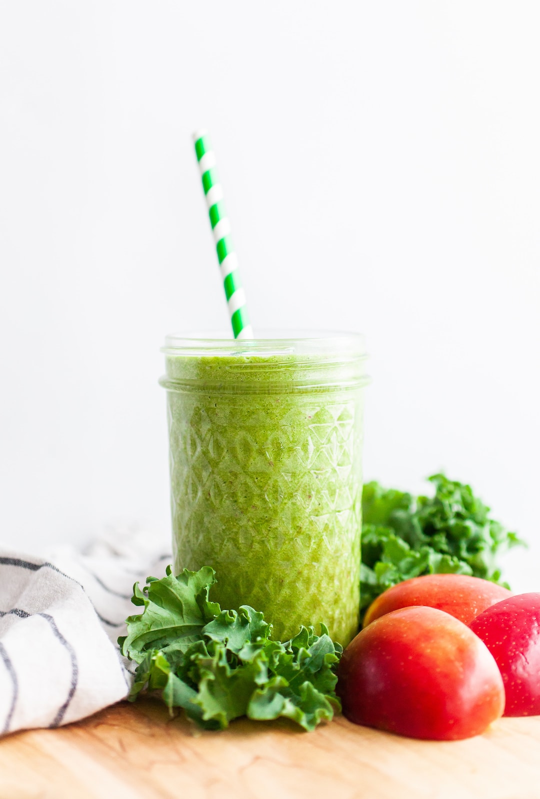 image of a green smoothie surrounded by kale and red apple with a green striped straw