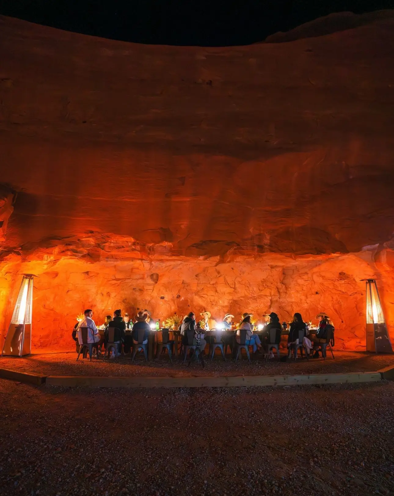 A cozy, intimate dinner gathering set against the dramatic red rock backdrop of a canyon cave. Warm lighting and rustic décor create an unforgettable desert dining experience at Moab, Utah.