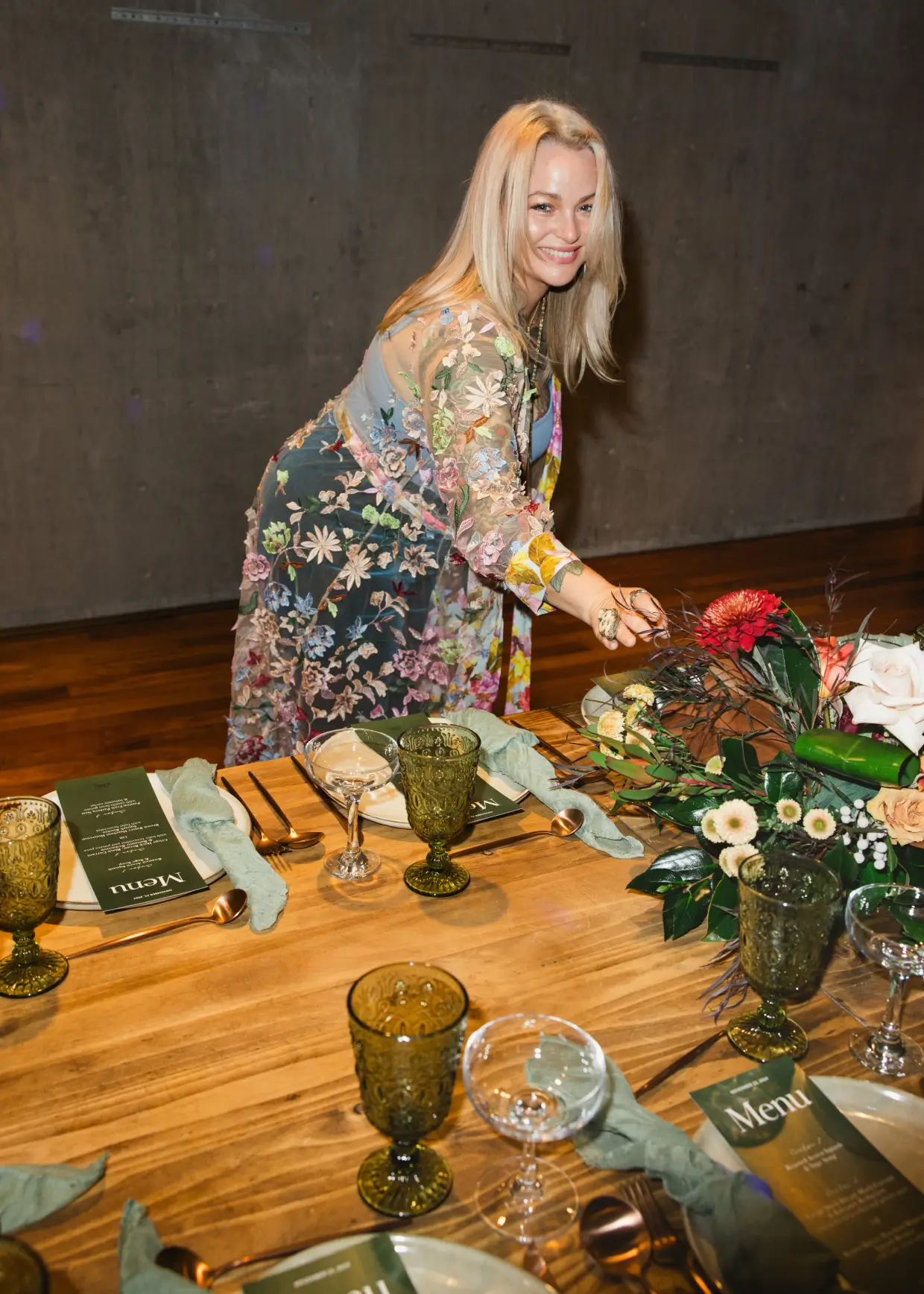 Chef Joann smiling beside a stylishly set table with elegant glassware and fresh green floral arrangements at her signature Client Soirée in La Jolla.
