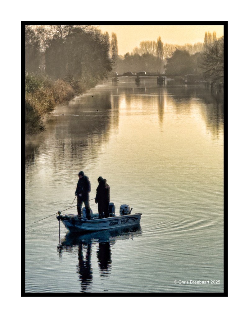 Two people fishing from a small boat on a calm river during an autumn sunrise, with trees reflecting in the water.