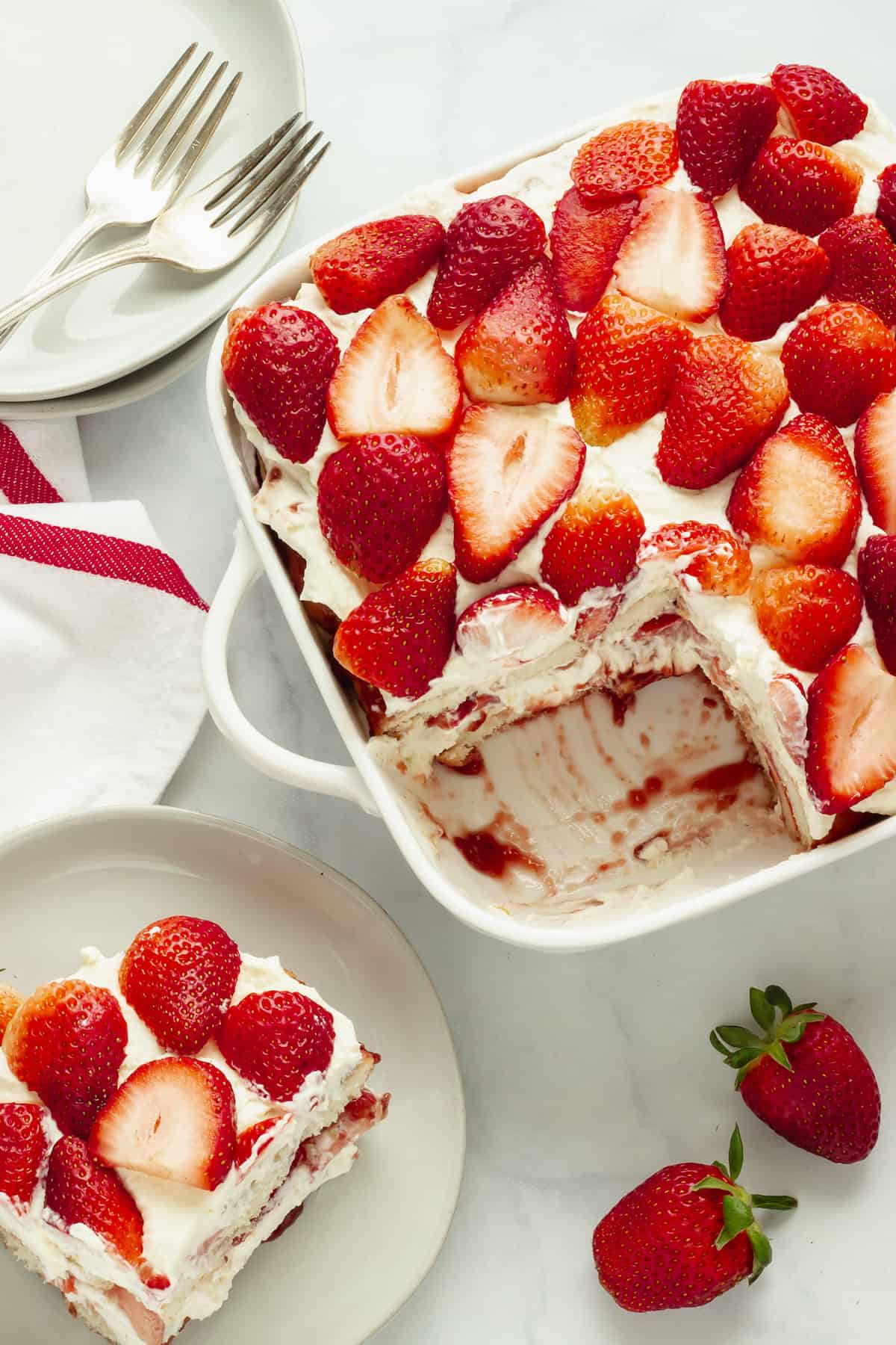 A white baking dish with strawberry tiramisu. A small plate with a serving of the dessert, a red and white striped napkin, and fresh strawberries are in the background.