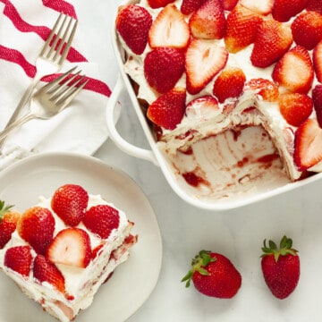 A white casserole dish with strawberry tiramisu and one serving plate with a piece of the dessesrt. Fresh strawberries, a red and white stripe napkin, and two forks are in the background.