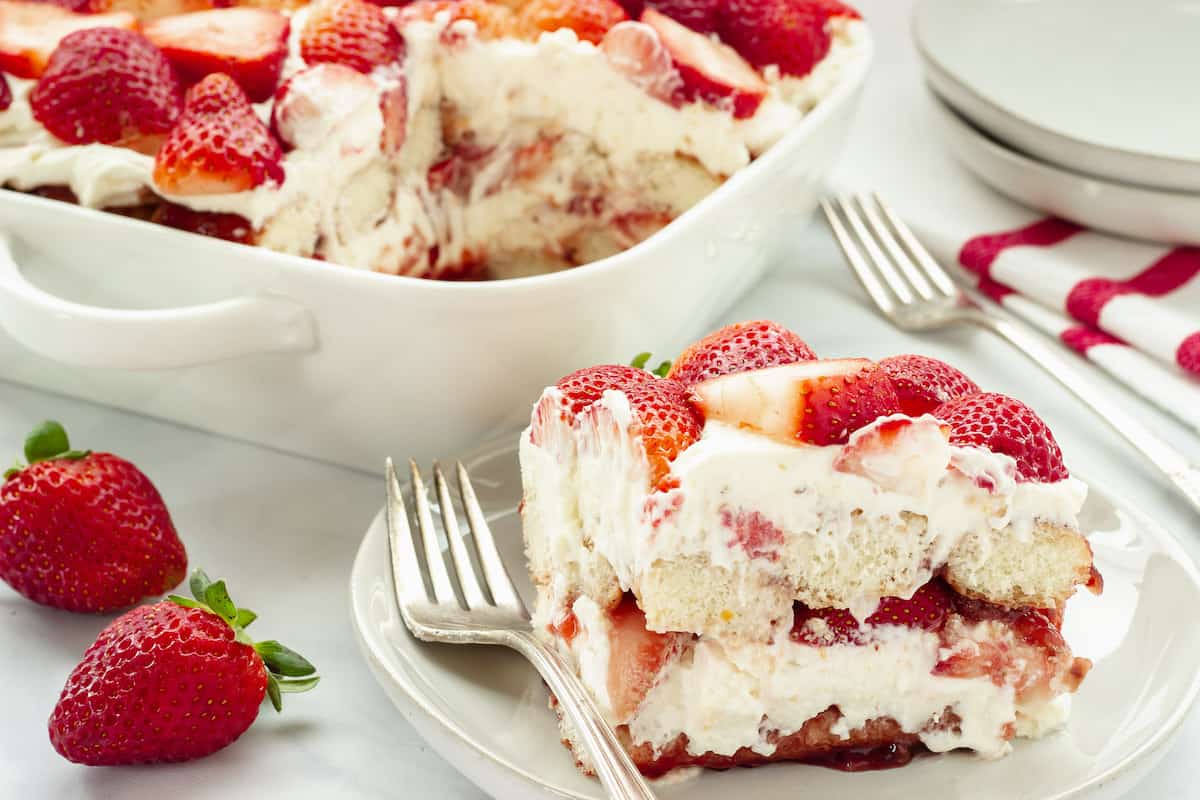 A white plate and fork with a slice of strawberry tiramisu. A pan of the dessert is in the background.