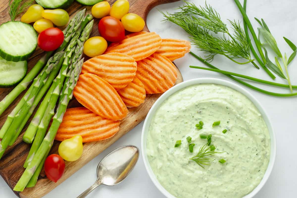 A bowl of green goddess dip next to a platter of fresh vegetables. Fresh herbs are in the background.