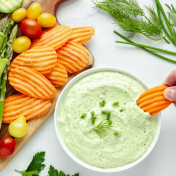 A hand dipping a carrot into a bowl of green goddess dip. A platter of vegetables is in the background.