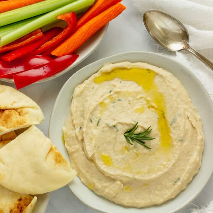 A plate spread with white bean dip. Cut vegetables and pita bread are in the background.