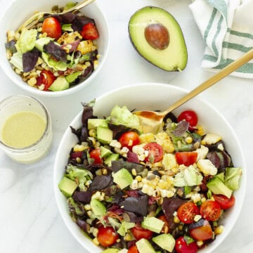 A large white serving bowl of BLT salad with a smaller portion off to the side. A jar of dressing and a cut avocado are in the background.