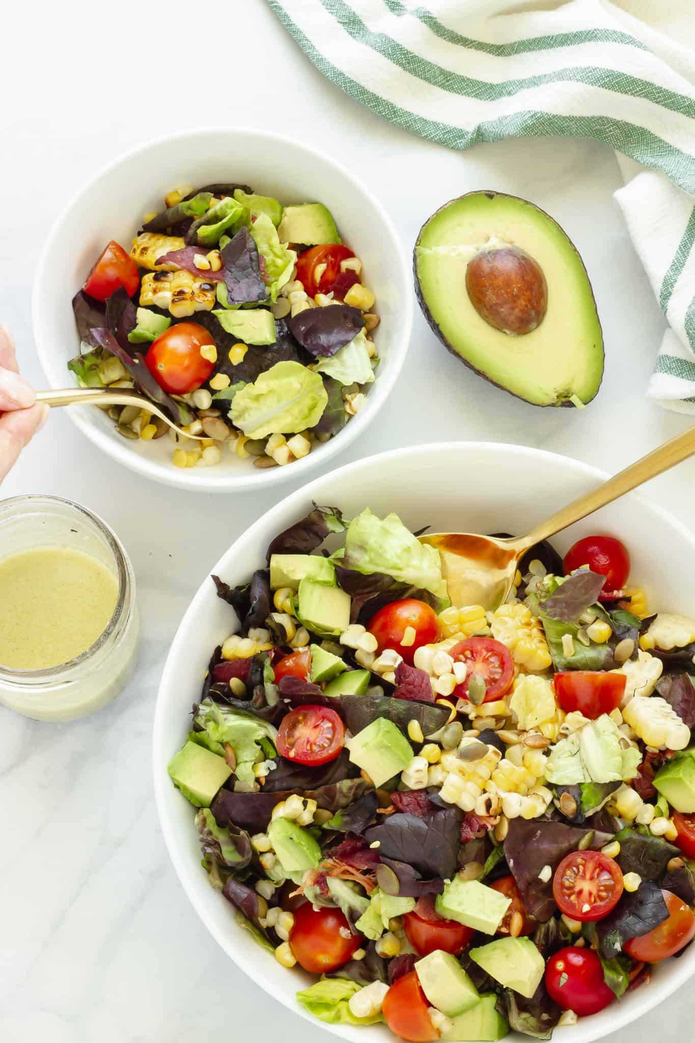 A large serving bowl of BLT salad with a smaller portion next to it. A hand is holding a fork in the smaller portion. A jar of dressing and a cut avocado are in the background.