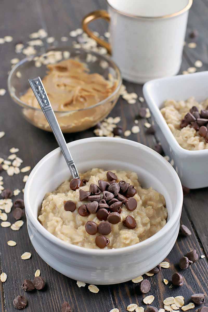 Vertical oblique shot of two white ceramic bowls, one round and one square, filled with oatmeal topped with chocolate chips, with a glass bowl of creamy peanut butter and a white mug with a gold handle in the background, and a spoon stuck into the bowl in the foreground, on a dark brown wood surface with scattered oats and dark chocolate.