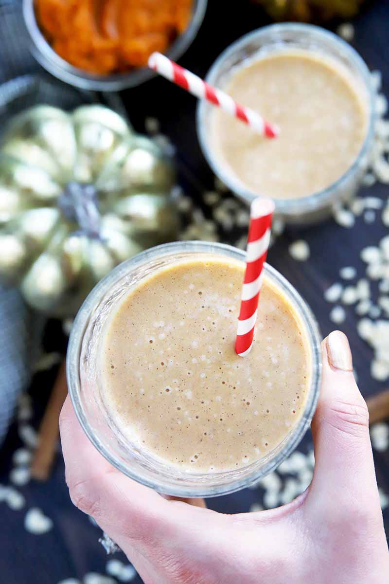 Vertical overhead image of a hand with champagne-colored painted nails holding a glass of pumpkin smoothie with a red and white striped paper straw sticking out of it, with another identical cup on the black surface below with scattered uncooked rolled oats, a gold decorative artificial pumpkin, and a small dish of orange squash puree, with a blue-gray cloth to the left of the frame.