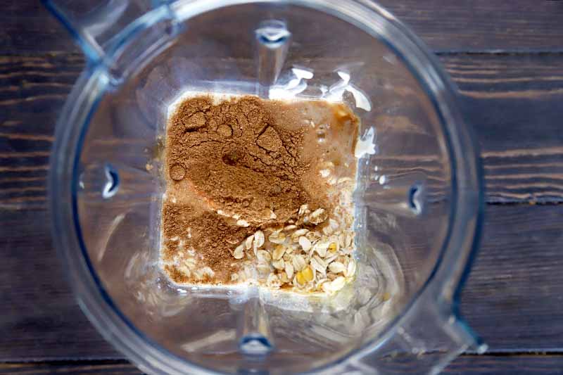 Horizontal image of a mixture of spices, oats, and dairy in a clear plastic high-speed blender pitcher, on a brown wood surface.
