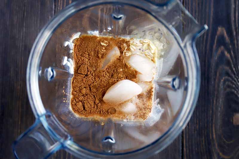 Horizontal overhead image of a mixture of ground spices, dairy, oats, and ice in a plastic blender pitcher, on a dark brown wood table.