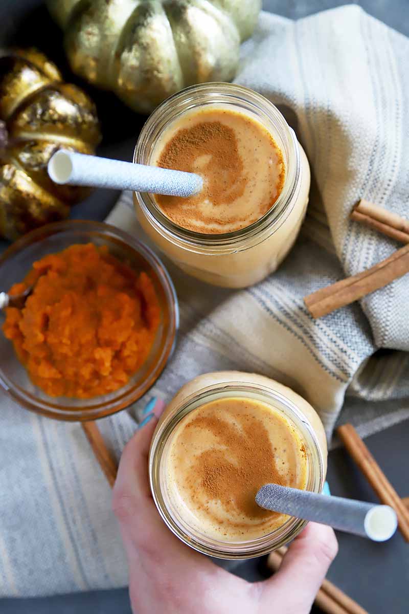 Vertical top-down image of a hand holding one of two light orange drinks in glass jars with straws, next to an orange puree and towel.
