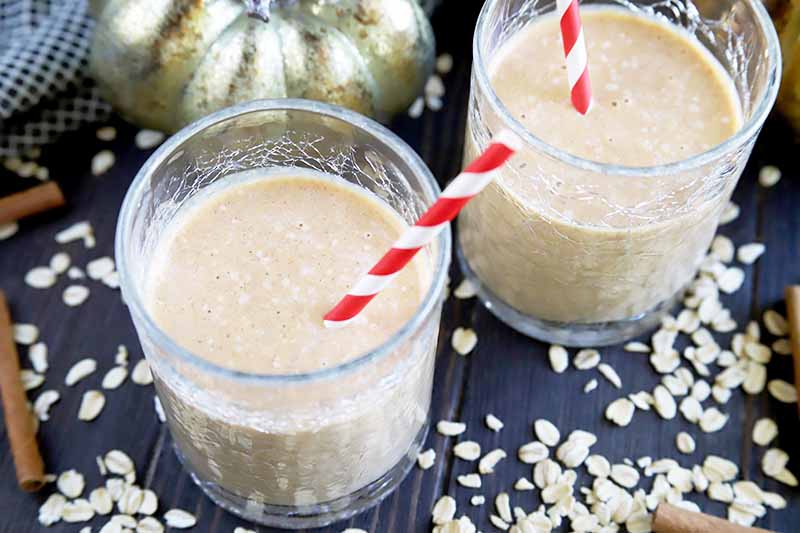 Horizontal oblique overhead image of two glasses of pumpkin smoothie with paper red and white striped straws, on a dark gray surface with scattered oat and cinnamon sticks, and a gold metallic decorative pumpkin.