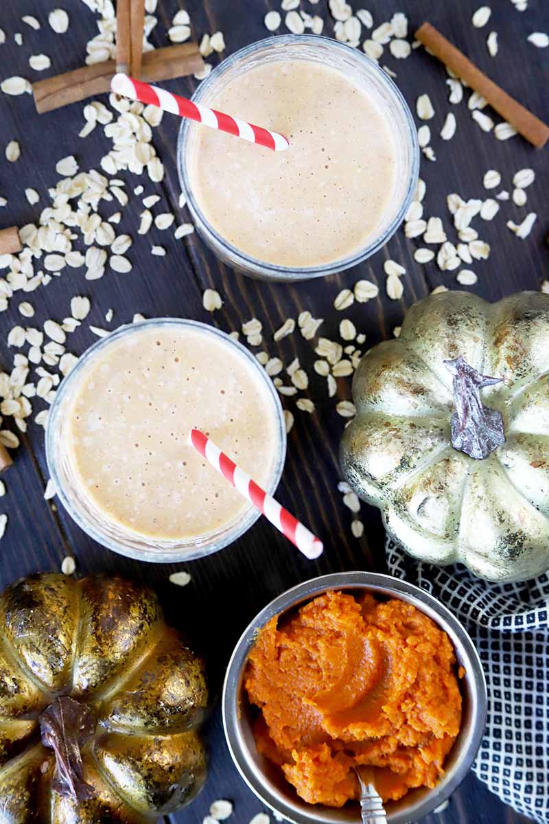 Vertical overhead image of two short glasses of pumpkin oatmeal smoothie with red and white paper straws, two gold metallic decorative pumpkins, a small bowl of orange squash puree, a gray-blue dish towel, and scattered uncooked rolled oats and cinnamon sticks, on a dark gray surface.