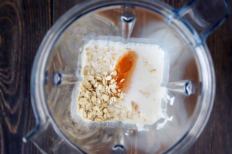 horizontal overhead image of orange pumpkin puree, uncooked rolled oats, and milk in the bottom of a clear plastic blender pitcher, on a dark brown wood surface.