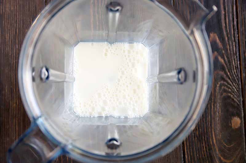 Horizontal overhead image of frothy milk in the bottom of a clear plastic pitcher on a dark brown wood surface.