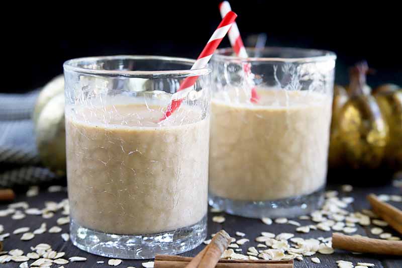 Horizontal image of two clear lowball glasses with a crackle surface, filled with a homemade pumpkin spice and oat smoothie, with red and white striped paper straws, on a dark gray surface with a gold decorative pumpkin, a blue-gray cloth, and scattered rolled oats and cinnamon sticks, against a black background.
