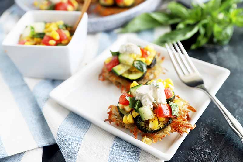 Horizontal image of a white plate with a potato fritter topped with sliced vegetables, a salsa, and a dollop of yogurt next fo a fork and another glass bowl filled with salsa.