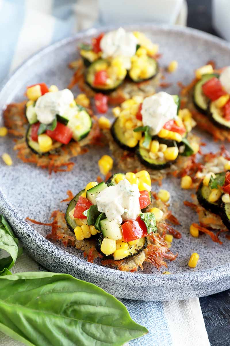 Vertical image of a gray plate with individual savory pancakes topped with assorted vegetables and a dollop of yogurt next to basil leaves.