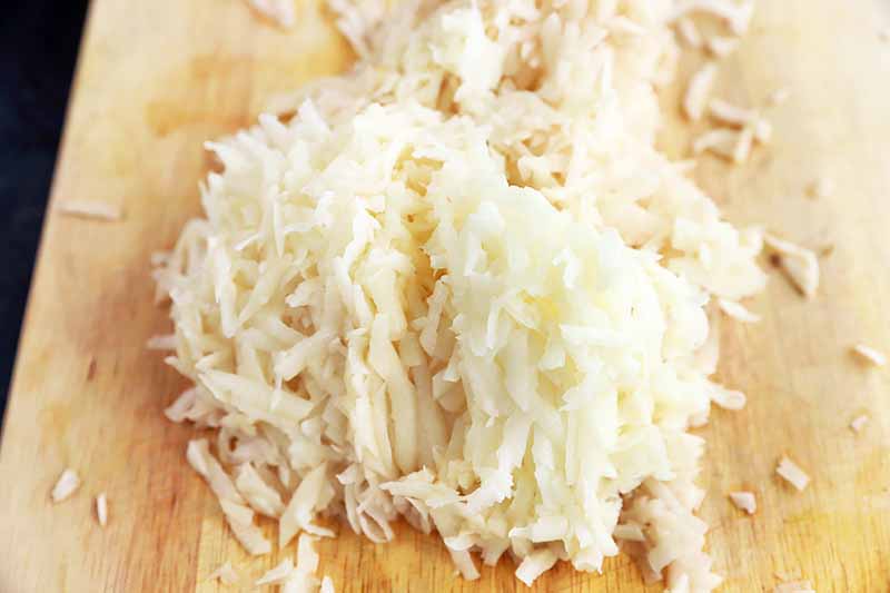 Horizontal image of grated potatoes on a wooden cutting board.