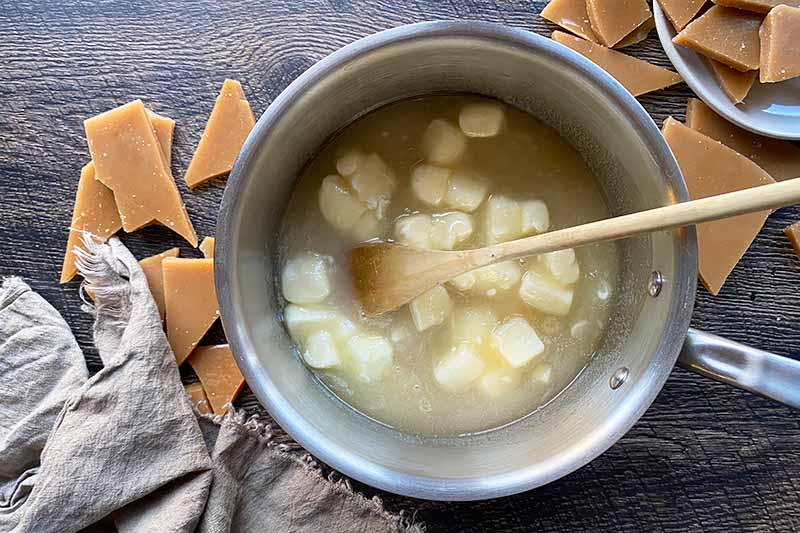 Horizontal image of melting butter cubes in a melted sugar mixture in a pot stirred by a spoon.