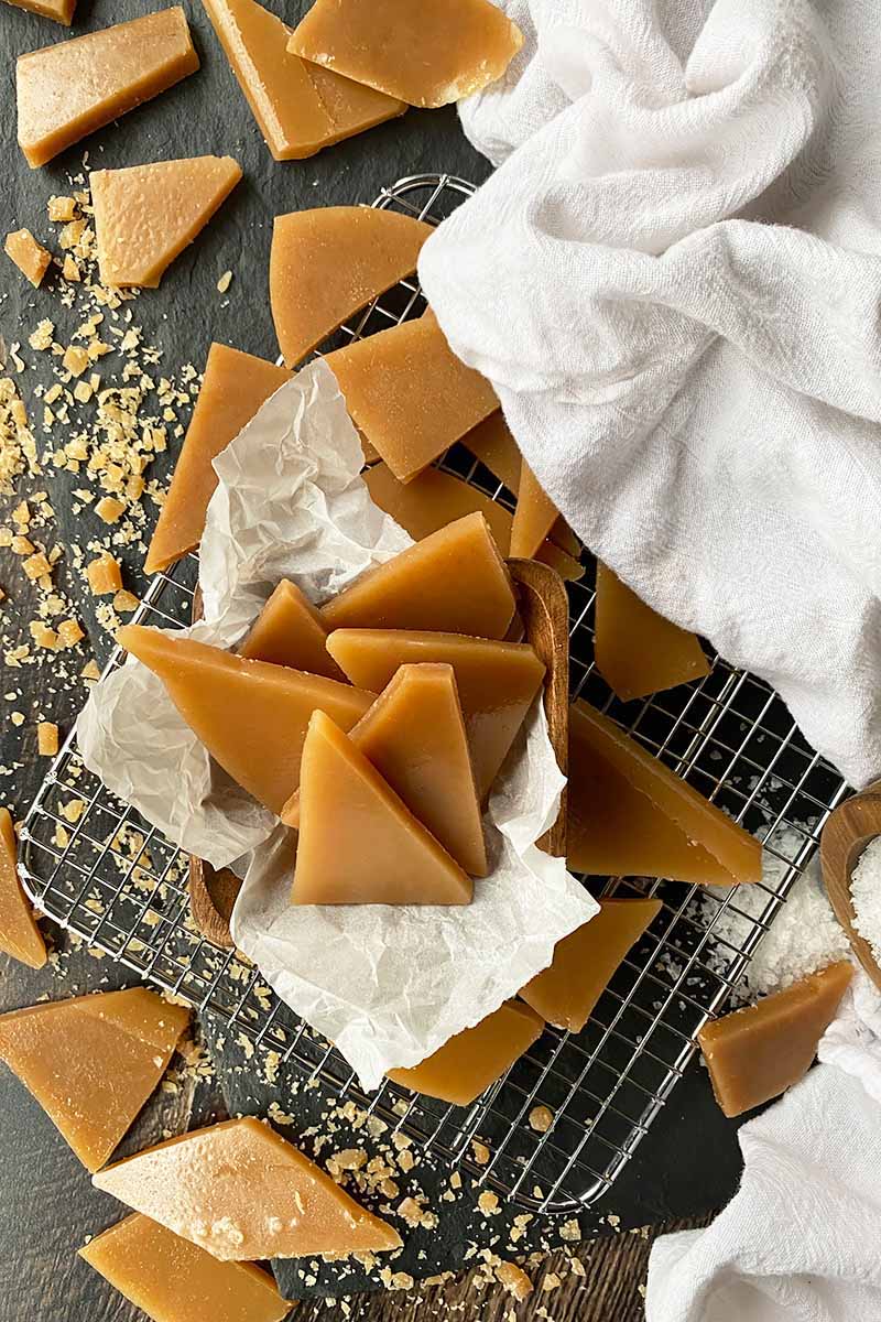 Vertical image of scattered toffee pieces on a dark surface with parchment paper, a white towel, and a cooling rack.