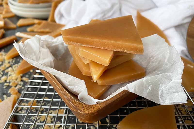 Horizontal image of a wooden bowl lined with parchment paper and filled with pieces of hard caramel candy on a cooling rack next to a white towel.
