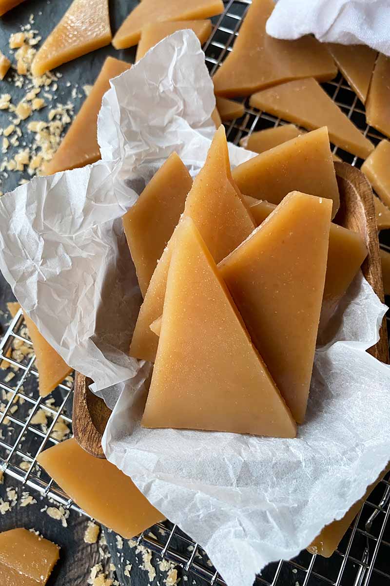 Vertical image of a wooden bowl lined with parchment paper filled with pieces of caramel candy on a cooling rack.