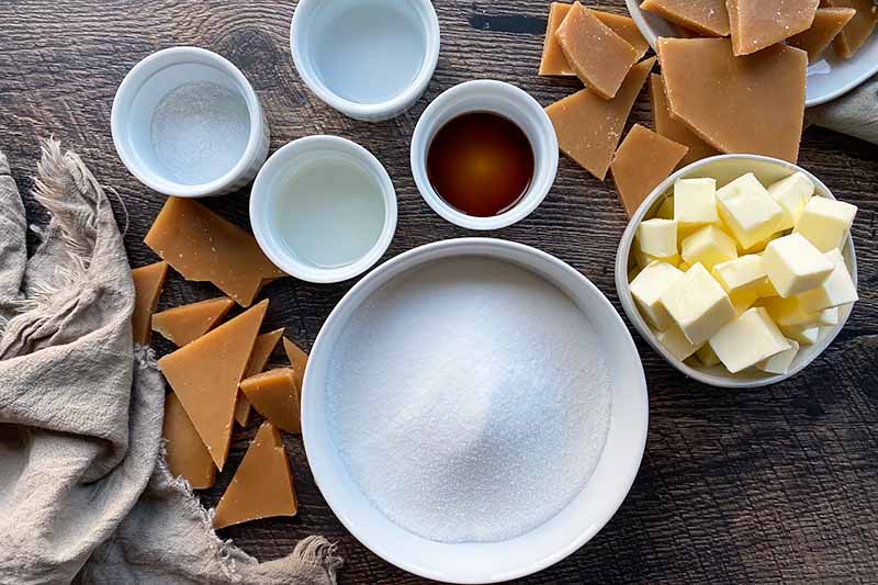 Horizontal image of assorted wet and dry ingredients in assorted white bowls next to toffee pieces and tan towels.