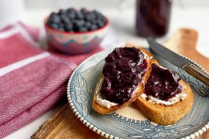 Horizontal image of slices of toast spread with butter and jelly next to a bowl of fruit and a red towel.