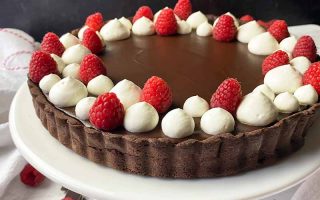 Horizontal image of a dessert with a cocoa crust garnished with whipped cream dollops and raspberries on a white cake stand.