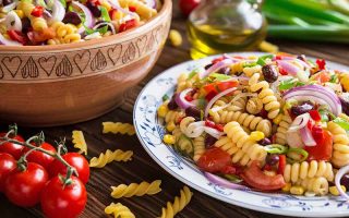 Horizontal image of a large bowl and a plate of a rotini and mixed vegetable and bean recipe next to a wooden spoon and cherry tomatoes.