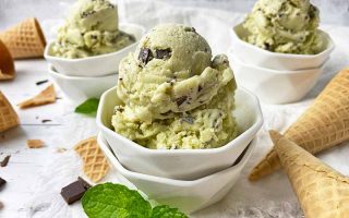 Horizontal close-up image of scoops of a light green ice cream in white bowls next to fresh herbs.