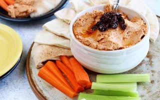 Horizontal image of a light red spread in a white bowl on a plate with celery, carrots, and pita wedges.