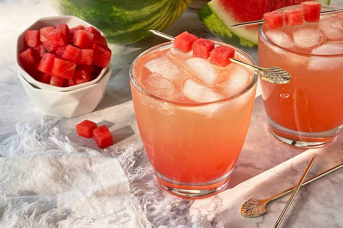 Horizontal image of glasses filled with a light pink beverage filled with ice with a garnish on a pick, next to a white bowl filled with cubes of fresh fruit.