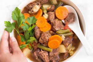 Horizontal image of a hand holding the side of a bowl full of a hearty meat and vegetable soup with a spoon.