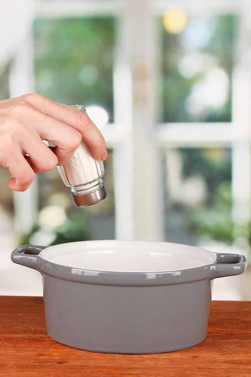 Vertical image of a hand seasoning food in a pot on a wooden table.