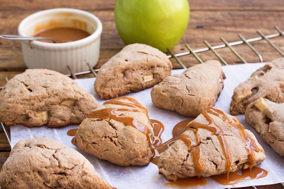 Horizontal image of apple scones, some drizzled with caramel sauce, on a cooling rack in front of whole fruit and a ramekin.