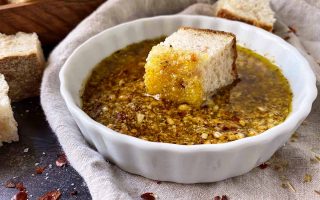 Horizontal image of a shallow white bowl, resting on a tan towel, filled with flavored dip and a cube of sourdough resting in it.