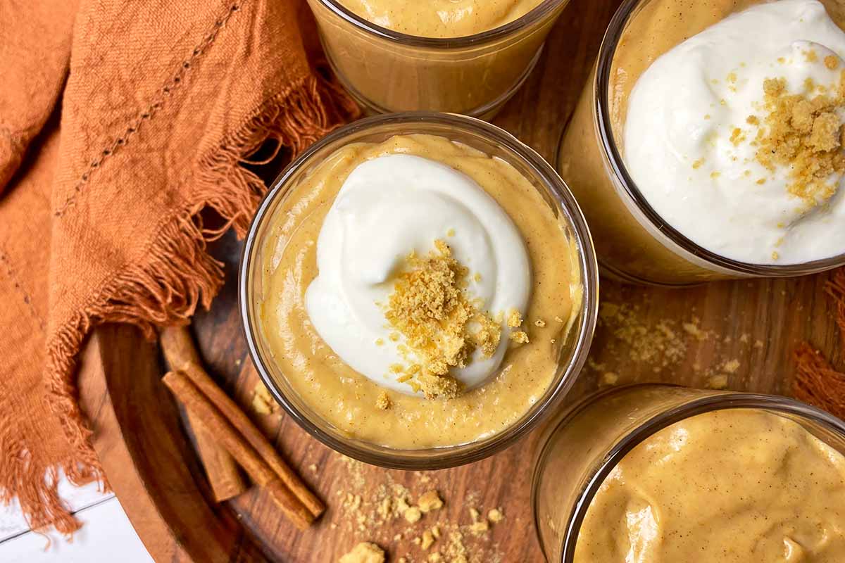 Horizontal top-down image of four glasses filled with a spiced creamy dessert on a wooden tray next to cinnamon sticks and orange towels.
