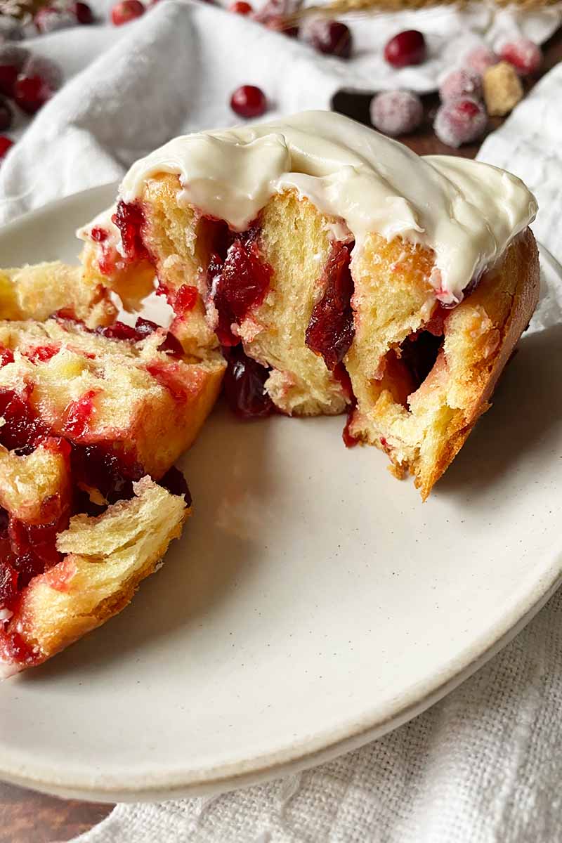 Vertical close-up image of a halved pastry on a white plate with a red fruit filling and white frosting on top.