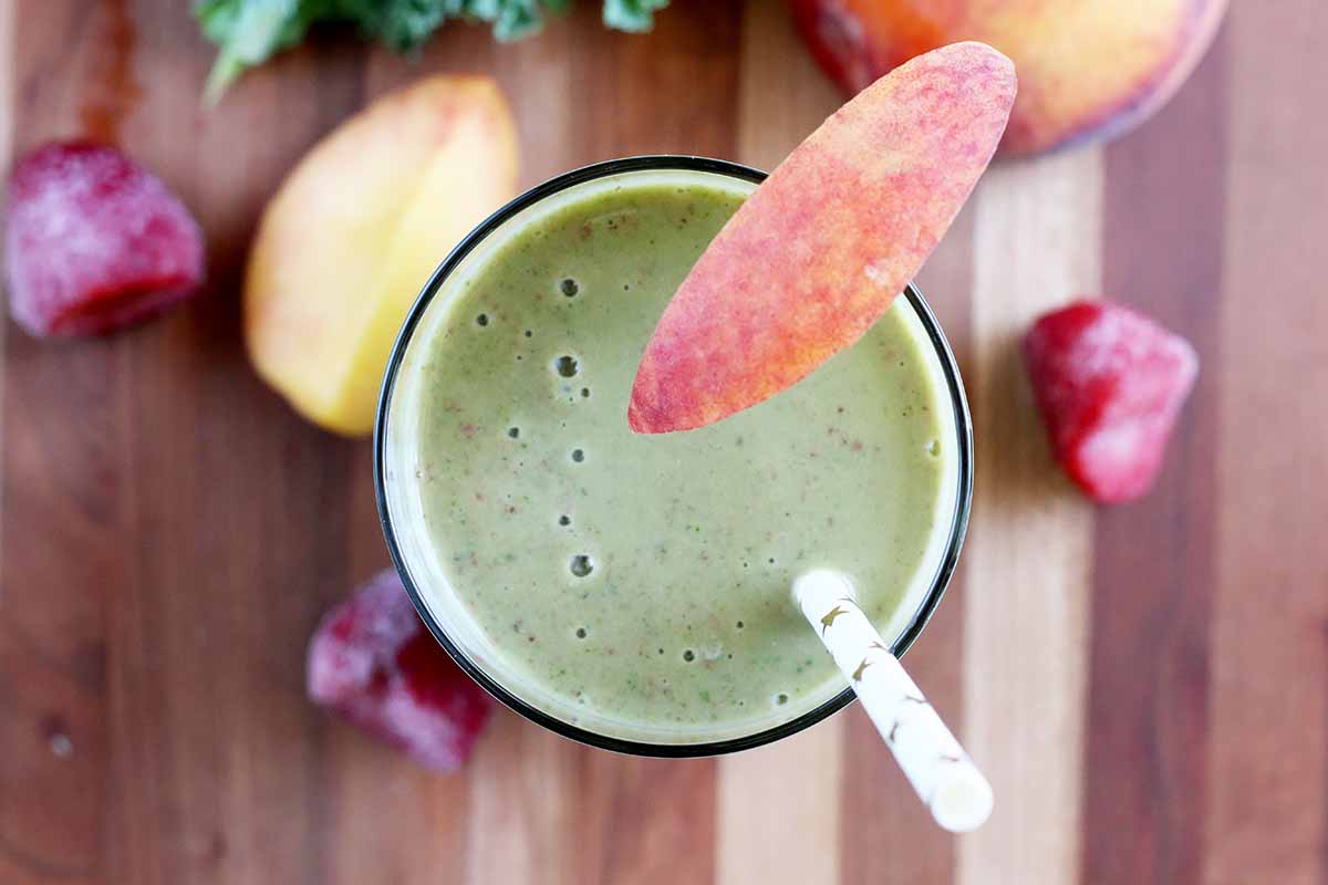 Horizontal image of a peaches and greens blended beverage in a cup with a peach garnish and a straw.