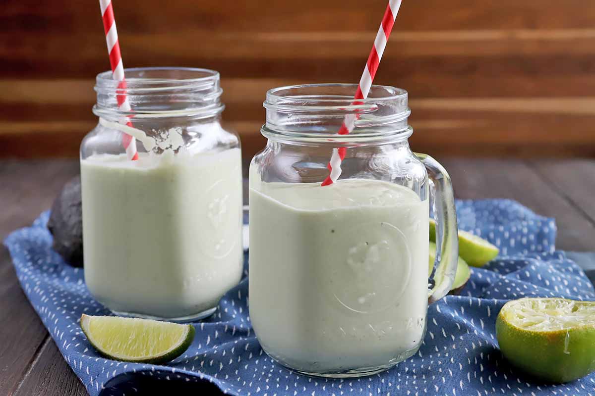 Horizontal image of two glasses filled with an icy avocado beverage on top of a blue towel next to limes.