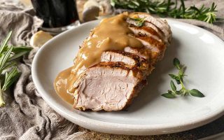 Horizontal image of shingled slices of seared meat topped with gravy on a white plate next to herbs and tan towels.