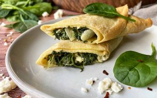 Horizontal image of a halved and stack thin pancake with a greens, onion, and cheese stuffing on a plate next to a wooden serving platter and green leaves.