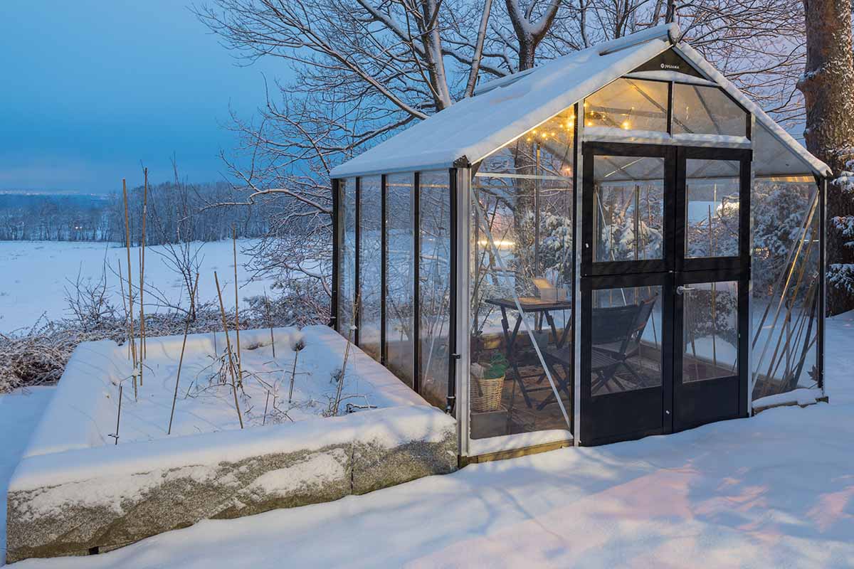 A close up horizontal image of a greenhouse in the backyard surrounded by snow set next to a raised bed garden.