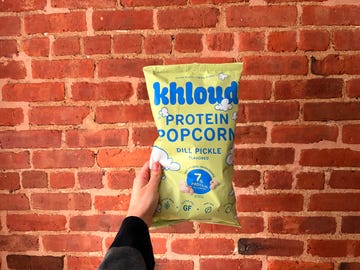 A hand holding a bag of protein popcorn against a brick wall.