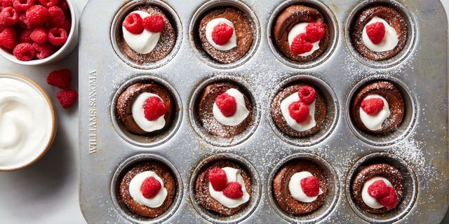 Mini Chocolate Dutch Baby Pancakes miniature chocolate flavored dutch baby pancakes sitting in the wells of a muffin tin and garnished with whipped cream, raspberries, and powdered sugar